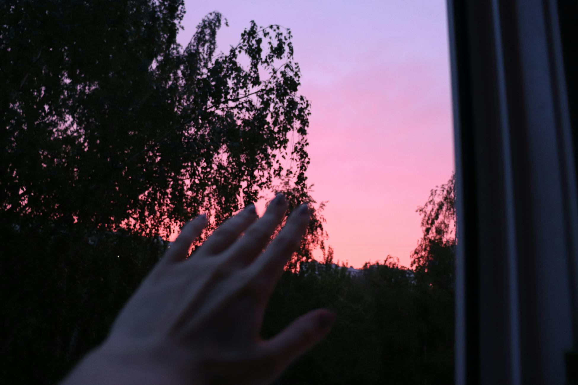 A hand reaching out to a stunning pink and purple sunset view from a window.