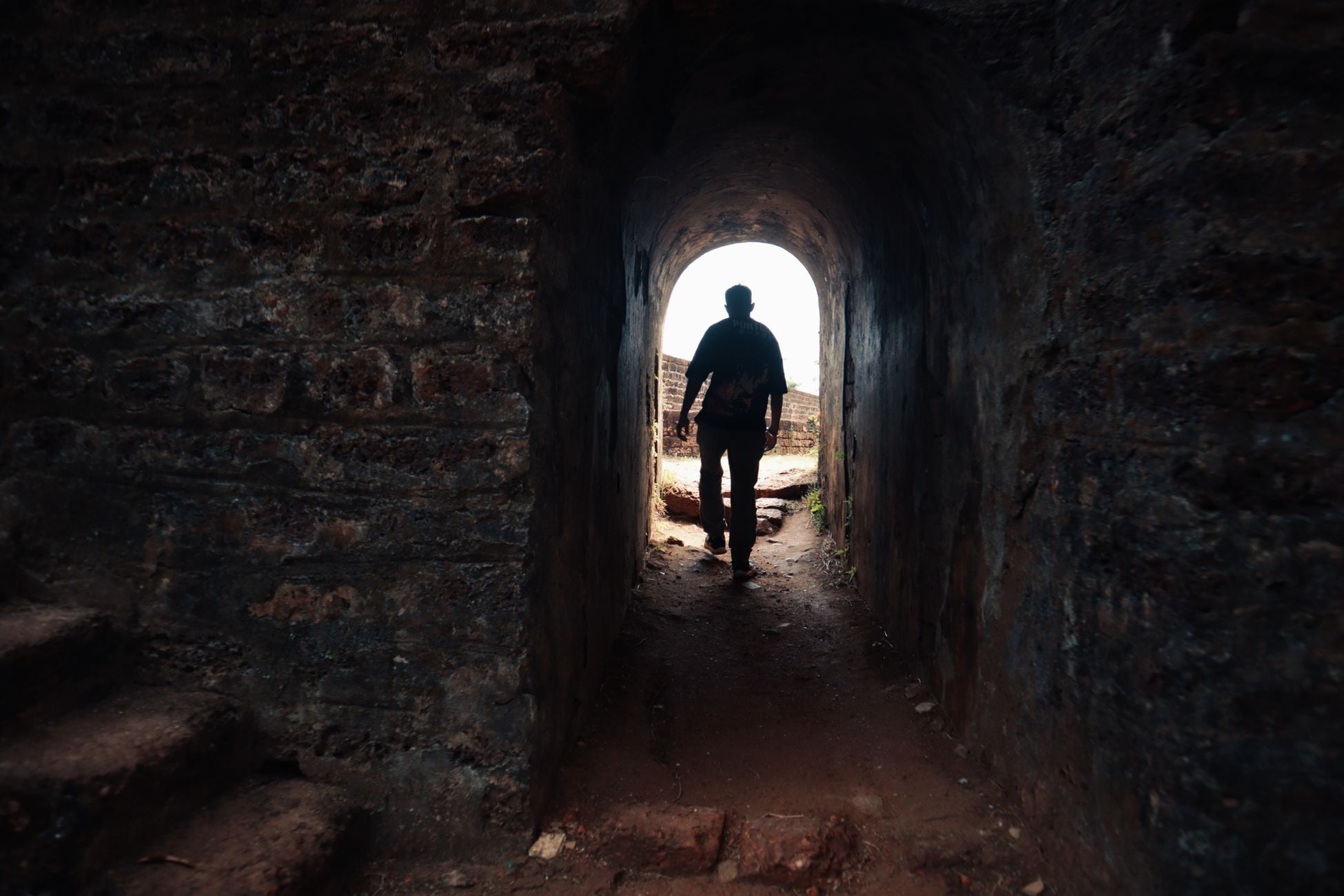 A solitary figure walks through a historic stone tunnel towards the light.
