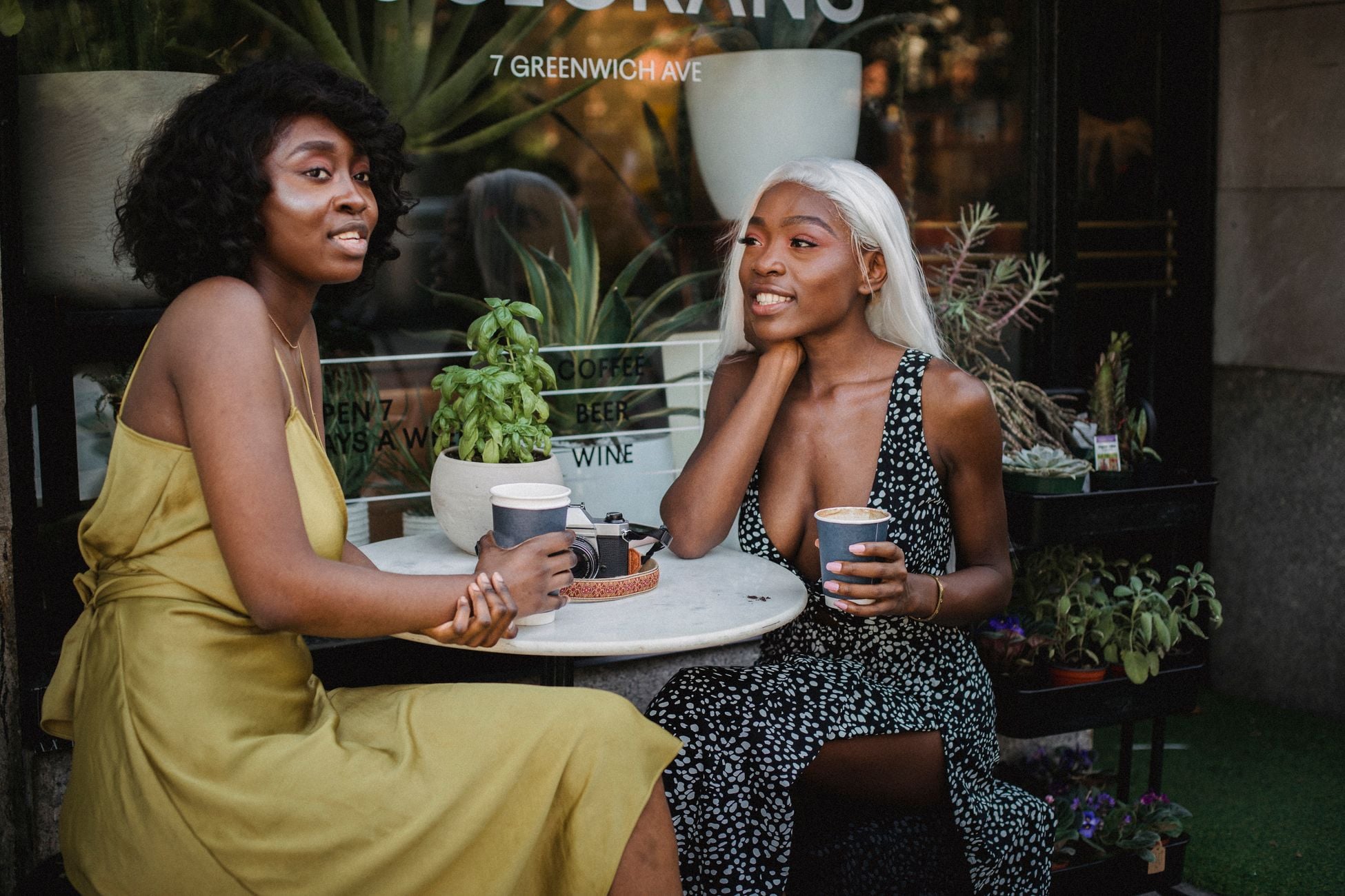 Two women enjoy coffee and conversation at a trendy café with vibrant plant décor.